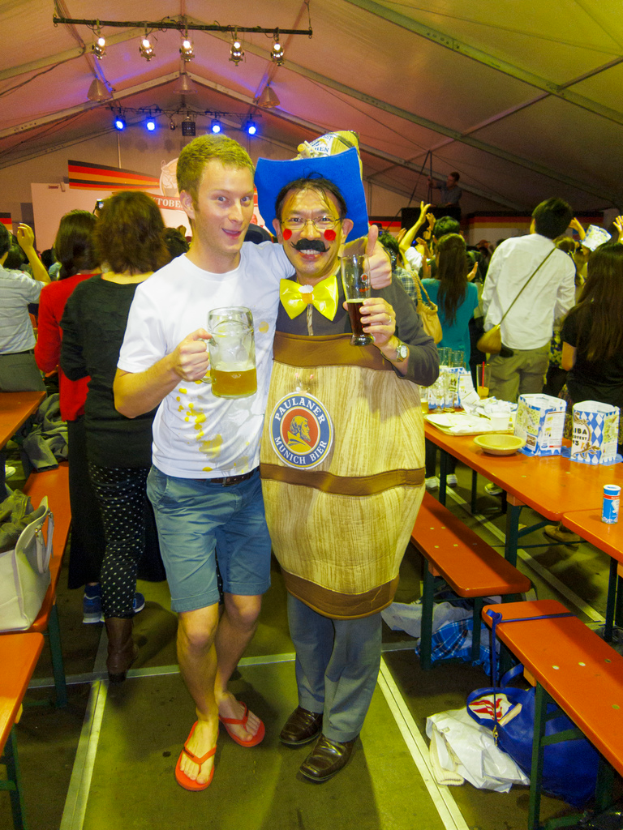 Zwei Personen stehen zusammen, eine hält ein Kind und ein Bier, die andere trägt ein Drama-Kostüm, mit einem nahen Tisch voller Essen und einer Gruppe von Menschen im Hintergrund.