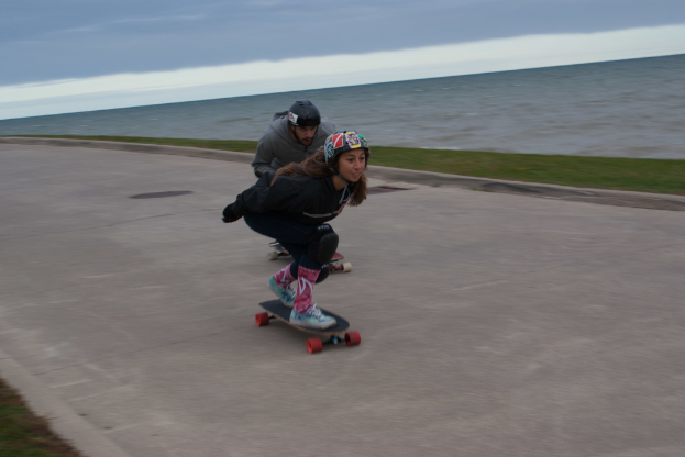 Zwei Personen mit Helmen fahren auf einem Weg neben einem Fluss, mit Gras und Himmel im Hintergrund.