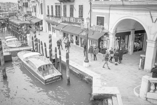 Ein Schwarz-Weiß-Bild eines Sees mit zwei Booten, die auf dem Wasser schwimmen, hölzernen Pfählen neben den Booten, Menschen, die auf dem Land gehen, und Gebäuden im Hintergrund.