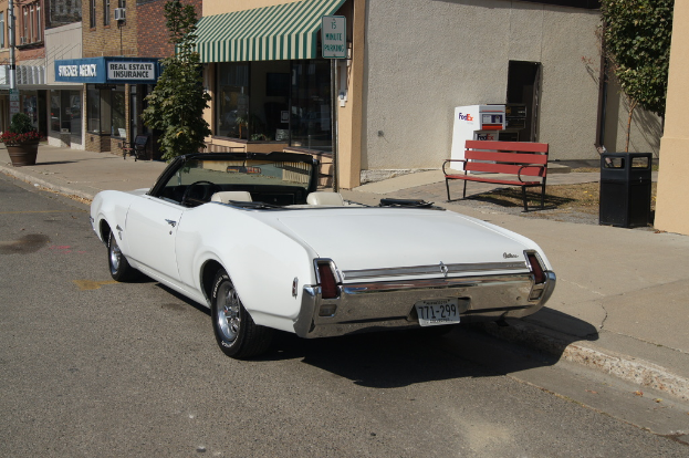 Ein weißes Auto mit der Nummer 771-299 steht auf der Straße, rechts daneben eine Bank und im Hintergrund ein Gebäude mit einem "Immobilienversicherung"-Geschäft.