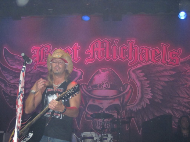 Eine Konzertszene mit einem Gitarristen links neben einem Mikrofon, einer Person in der unteren rechten Ecke, einem Banner im Hintergrund und Drogen in der Mitte unten, mit Bühnenbeleuchtung oben.