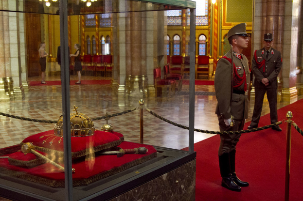 Fünf Personen stehen auf Teppichen mit Stangen, Seilen und Gegenständen in einer Glasbox zu ihrer linken, mit einer Säule, Licht, Stühlen und Kronleuchtern dahinter.