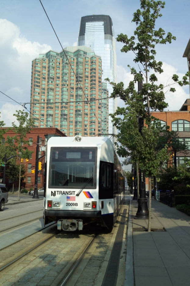 Ein Zug auf Schienen mit elektrischen Kabeln oben, umgeben von Bäumen, Polen und Gebäuden, mit einem Wolkenkratzer und Himmel im Hintergrund.