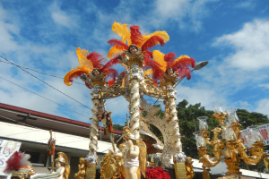 Ein Karnevalswagen mit goldenen und roten Federn, der Menschen trägt, wird in einem Umzug mit einem Gebäude, Bäumen, Drähten und einer Fahne in der Nähe, unter einem sichtbaren Himmel gezeigt.