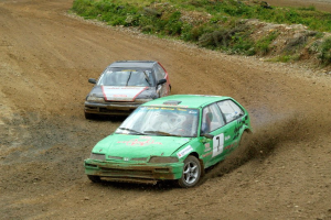 Zwei Autos mit Text und Zahlen fahren eine Schotterstraße entlang, umgeben von Gras, Pflanzen und Steinen, was auf ein Rallye-Veranstaltung hinweist.