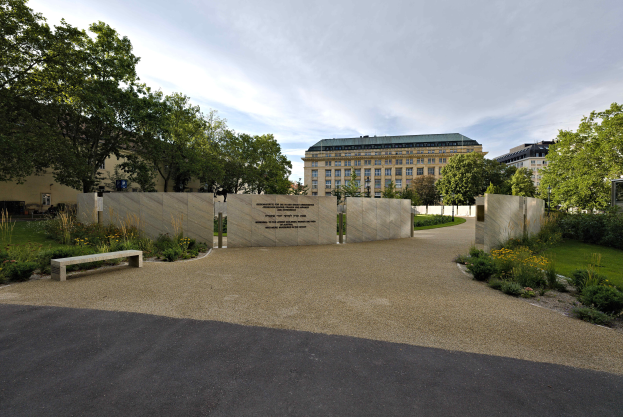 Eine Parkbank umgeben von Bäumen und Pflanzen, mit Gebäuden und einem bewölkten Himmel im Hintergrund und Text an der Wand hinter der Bank, der wahrscheinlich auf das Holocaust-Mahnmal in Berlin Bezug nimmt.