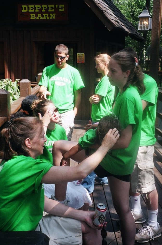 Mehrere Personen in grünen T-Shirts, einige sitzend und einige stehend, vor einem hölzernen Zaun mit einer Hütte, Bäumen und einer Lampe im Hintergrund.