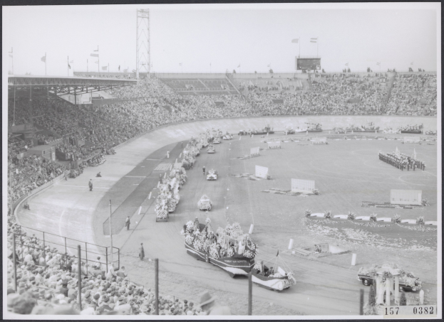 Schwarzes und weißes Foto einer Rennstrecke mit Zuschauern und Fahrzeugen, das Tribünen, Pfosten, Flaggen und den Himmel im Hintergrund zeigt.