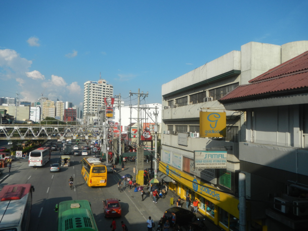 Eine belebte Stadtstraße mit verschiedenen Fahrzeugen, Fußgängern auf den Gehwegen, Gebäuden mit Schildern, Strommasten, eine entfernte Brücke, Bäume und ein bewölkter Himmel.