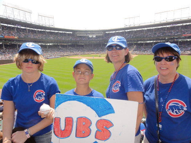 Drei Frauen in Kappen und Brillen, ein Junge mit Brille und Kappe, der einen Ball und ein Schild hält, stehen in einem Stadion mit Publikum und Feld dahinter, unter einem Himmel mit einigen Lichtern.