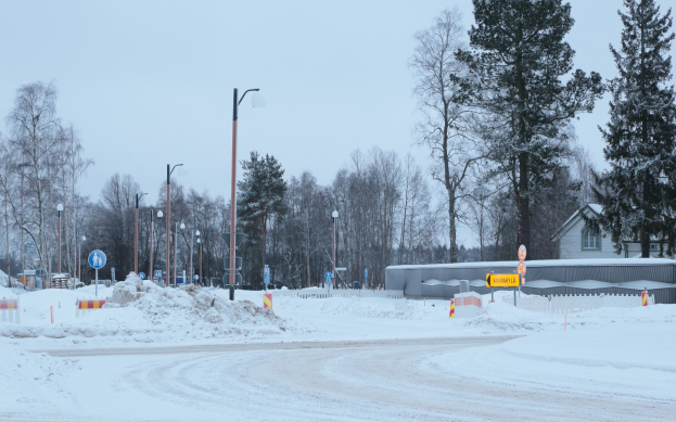 Eine verschneite Straße mit Laternen, Verkehrsschildern, Pfählen, Bäumen, einem Haus mit Dach und Fenstern und einem bewölkten Himmel.