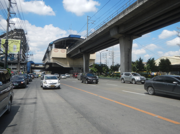 Vielbefahrene Hauptstraße mit fahrenden Autos, eine Brücke im Hintergrund, Strommasten mit Drähten, Bäume, Gebäude und ein bewölkter Himmel.