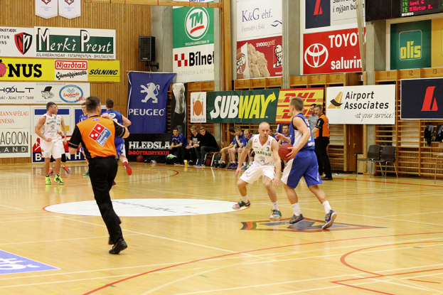 Ein Basketballfeld mit Spielern im Vordergrund und einer holzgetäfelten Wand mit Postern und Bannern im Hintergrund.