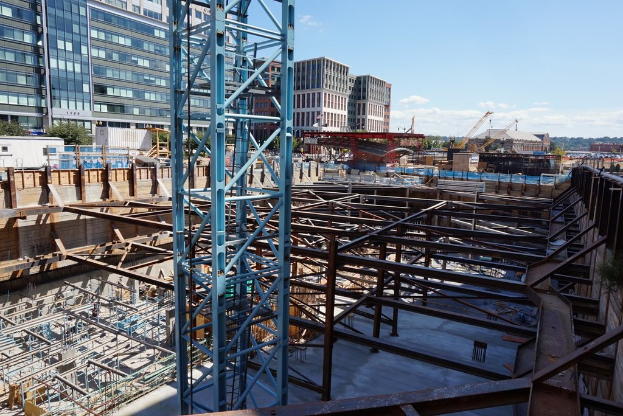 Das Bild zeigt eine Baustelle mit Gebäuden und Büros links, Eisenstangen und Maschinen rechts und verschiedenen Geräten und Kränen in der Mitte, unter einem Himmel in der rechten oberen Ecke.