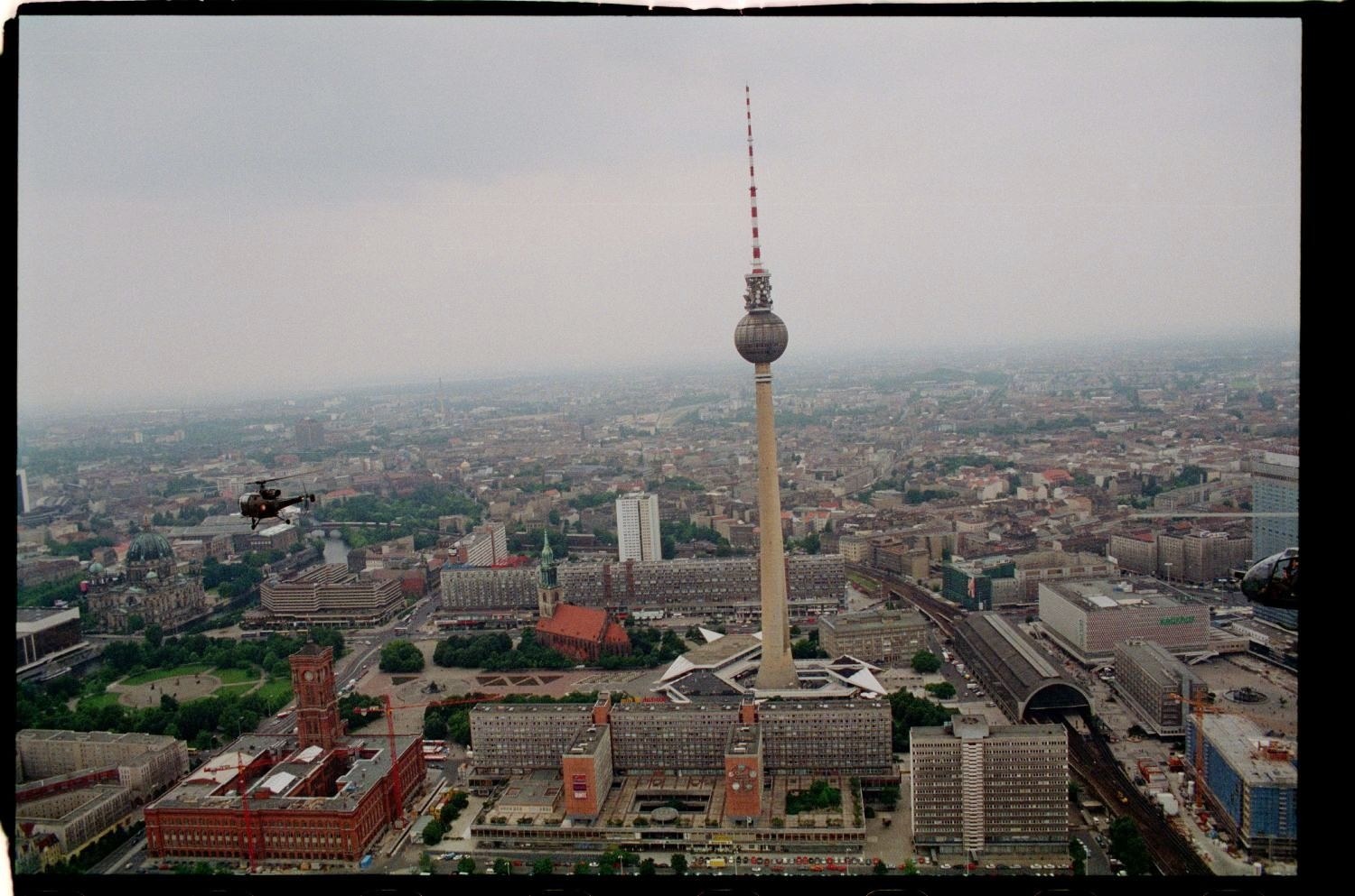 Luftaufnahme von Berlin, Deutschland, mit dem Fernsehturm in der Mitte und umgeben von Gebäuden, Bäumen, Straßen, Fahrzeugen und einem Hubschrauber unter einem klaren blauen Himmel.
