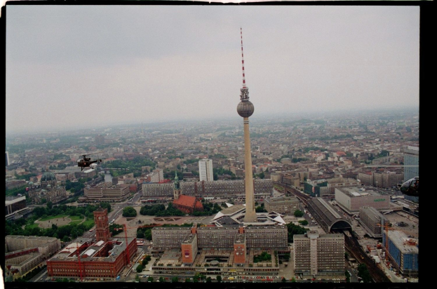Luftaufnahme von Berlin, Deutschland, mit dem Fernsehturm in der Mitte und umgeben von Gebäuden, Bäumen, Straßen, Fahrzeugen und einem Hubschrauber unter einem klaren blauen Himmel.