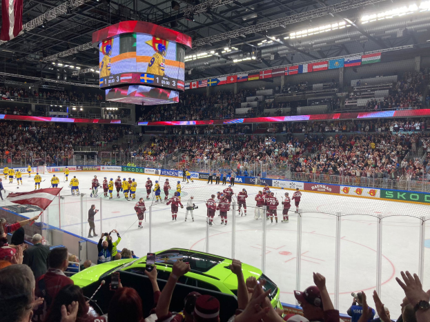 Ein Eishockeyspiel wird in einer großen Arena vor einem Publikum gespielt, einige halten Fahnen und ein Auto steht davor. Die Eisfläche ist von einem Zaun umgeben und Banner mit Texten hängen an der Decke, während die Decke mit Lichtern, einem Bildschirm und hängenden Fahnen geschmückt ist.