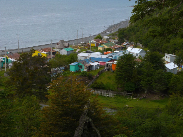 Ein Blick auf Häuser, eine Straße, Pfähle, einen Zaun, Bäume, Gras und einen Gewässerteil.
