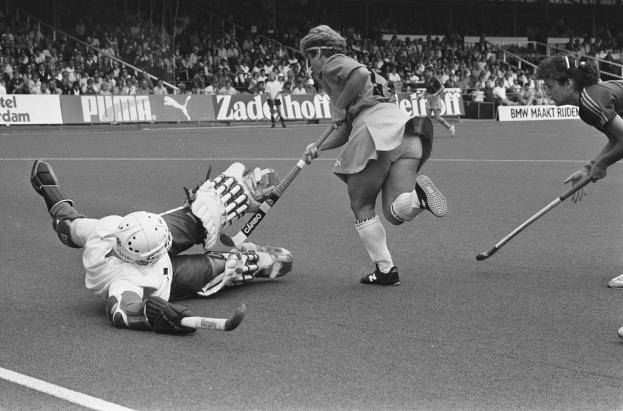 Schwarzes Bild von Frauen, die Feldhockey in einem Stadion spielen, mit Zuschauern auf den Tribünen, die Helme tragen und Hockey-Schläger halten, umgeben von Tafeln mit Text.