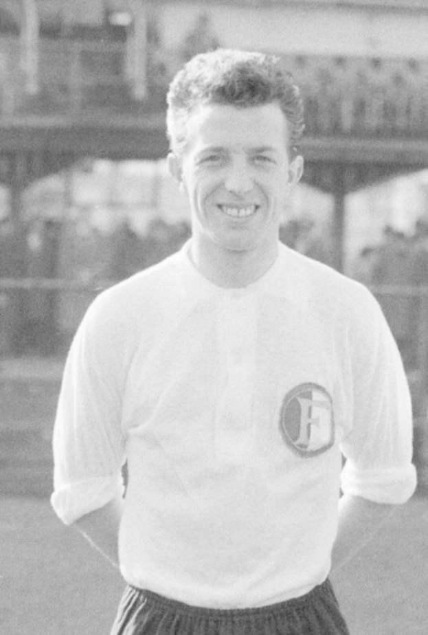 Ein Schwarz-Weiß-Foto eines grinsenden Mannes in einem T-Shirt und Shorts auf einem Fußballfeld mit einem Gebäude im Hintergrund, das mutmaßlich Mario Mandžukić ist.