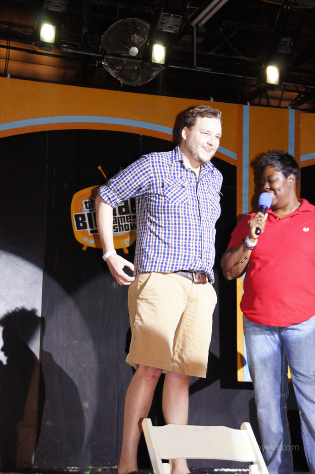 Eine Person in einem roten T-Shirt hält ein Mikrofon und spricht, während eine andere Person neben ihnen steht und lächelt; Fans und Lichter sind oben im Bild zu sehen.