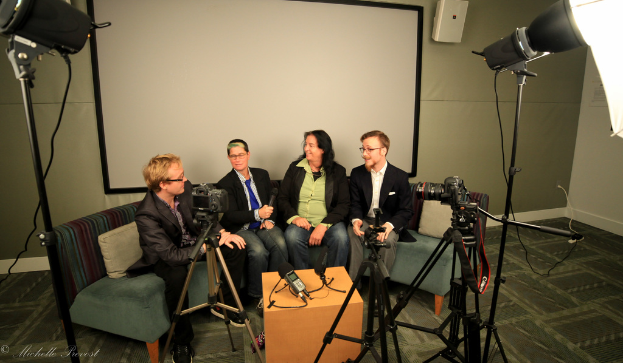 Eine Gruppe von drei Personen sitzt auf einem Sofa und gibt ein Interview. Links sitzt eine Person in einem schwarzen Mantel und Hose, in der Mitte eine Person in einem Mantel und einer blauen Krawatte mit einem Mikrofon in der Hand und rechts eine strahlende Person. Auf dem Tisch in der Mitte steht ein Mikrofonrekorder, Kameras befinden sich rechts und links und große Scheinwerfer sind vorhanden, sowie eine Projektionswand im Hintergrund.