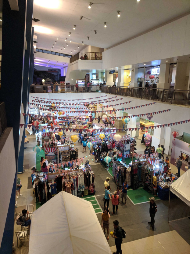 Ein volles Fest-Einkaufszentrum mit Ständen, hängenden Kleidern, dekorativen Fahnen, Ballons, sitzenden und stehenden Menschen, Geländern, Säulen und Deckenleuchten, ähnlich einem Weihnachtsmarkt im Mall of Asia.