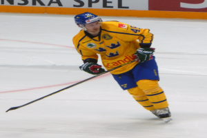 Ein Eishockeyspieler in Aktion auf dem Eis, der einen Helm, Handschuhe und Schlittschuhe trägt und einen Hockeystock hält, mit einer Tafel mit Text im Hintergrund.