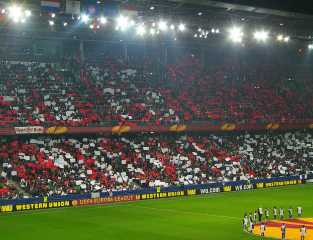 Ein Fußballspiel wird in einem großen, hell erleuchteten Stadion mit Zuschauern auf den Rängen und Menschen auf dem Feld im Vordergrund gespielt.