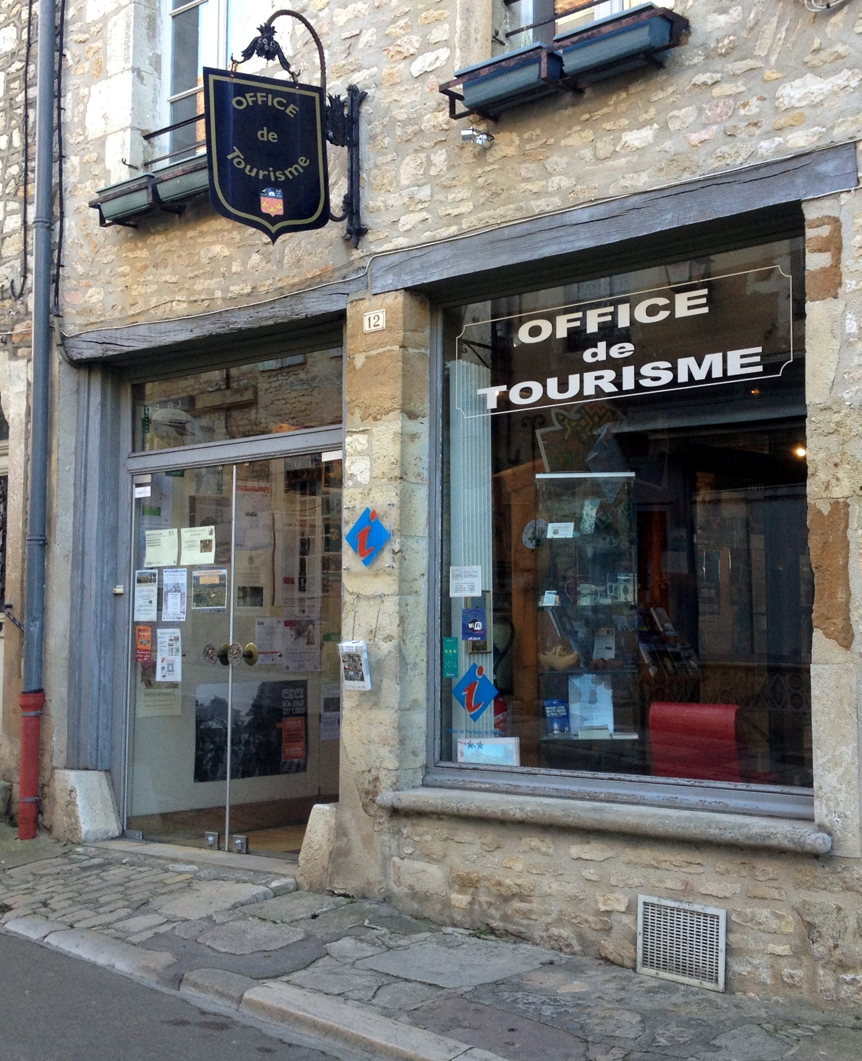 Ein Gebäude mit einem Schild, das "Office de Tourisme" lautet, Fenstern, einer Tür mit aufgeklebten Papieren, einem Rohr an der Seite und einer Straße unten.