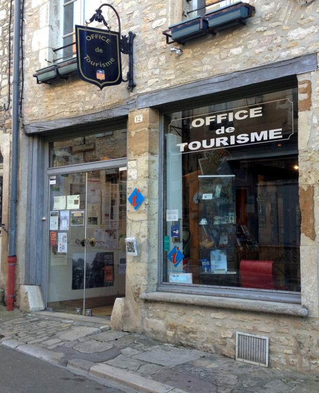 Ein Gebäude mit einem Schild, das "Office de Tourisme" lautet, Fenstern, einer Tür mit aufgeklebten Papieren, einem Rohr an der Seite und einer Straße unten.