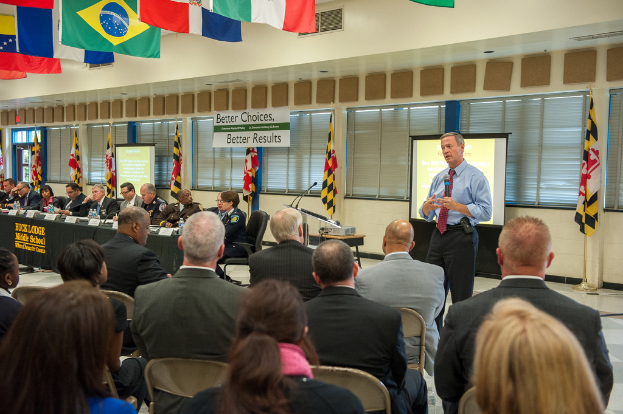 Eine Gruppe von Menschen, die auf Stühlen sitzen und einem Redner mit Mikrofon zugewandt sind, mit einem Projektionsschirm hinter ihm, einem Projektor auf einem Tisch, einer Personentafel mit Tisch, Wasserflaschen, Fahnenmasten, hängenden Fahnen, abgedeckten Fenstern und einer Tür auf der linken Seite.