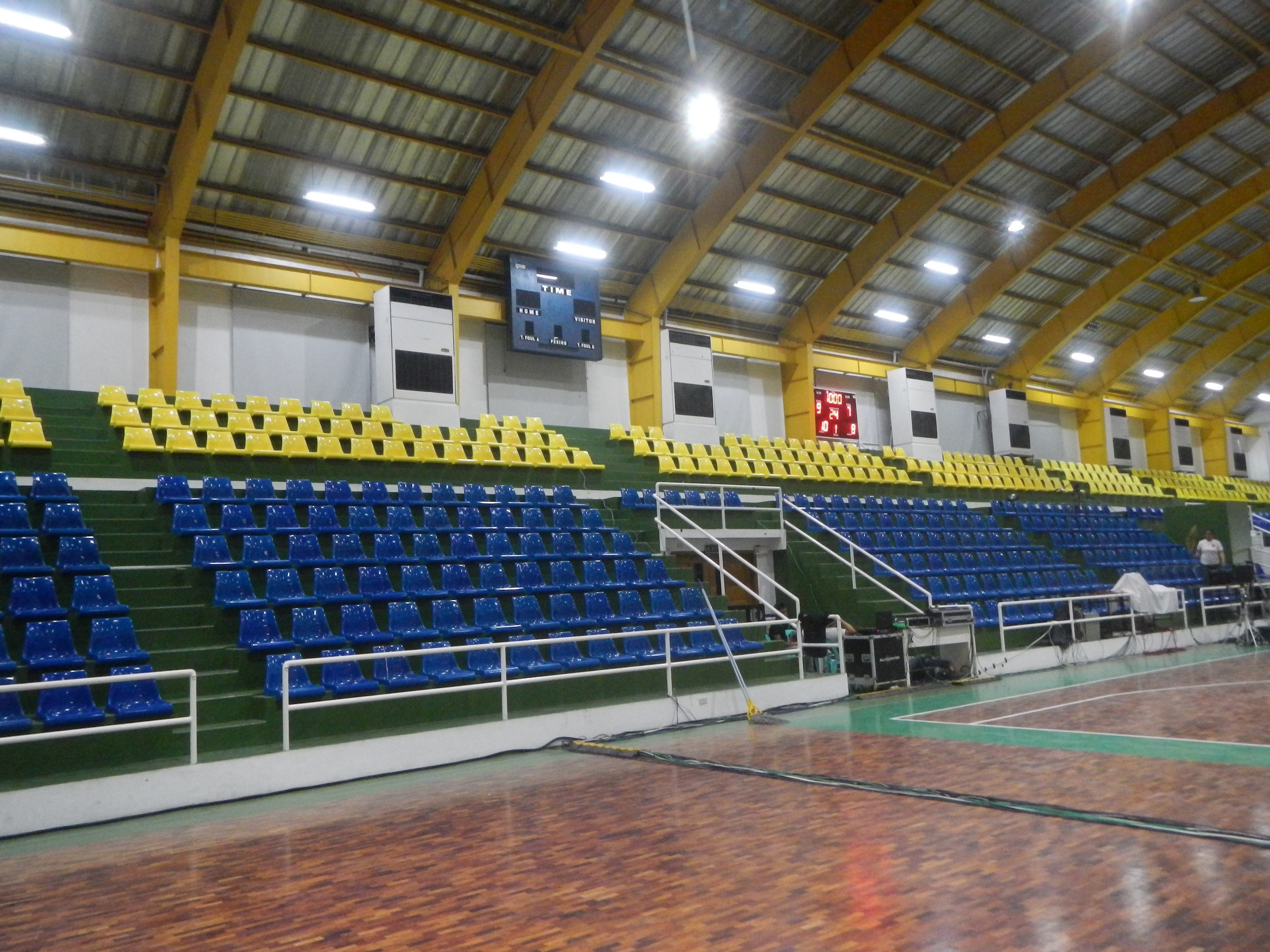 Großer Hallensportplatz mit Holzboden, blauen und gelben Sitzplätzen, Geländern, Treppen zum Sitzbereich, Wandbildschirmen, Deckenleuchten und einigen anwesenden Personen.