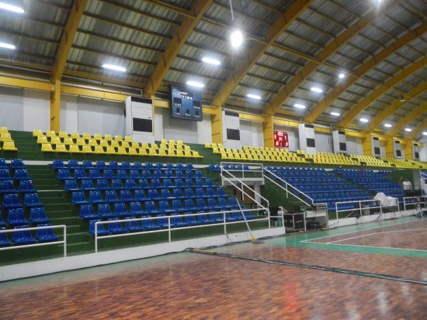 Großer Hallensportplatz mit Holzboden, blauen und gelben Sitzplätzen, Geländern, Treppen zum Sitzbereich, Wandbildschirmen, Deckenleuchten und einigen anwesenden Personen.