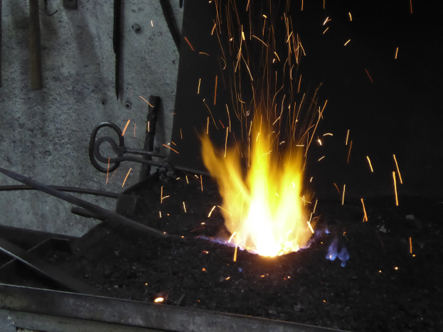 Ein Schmied formt Metall mit funkensprühenden Funken, Metallstangen und eine Wand links und ein Feuer auf der rechten Seite.