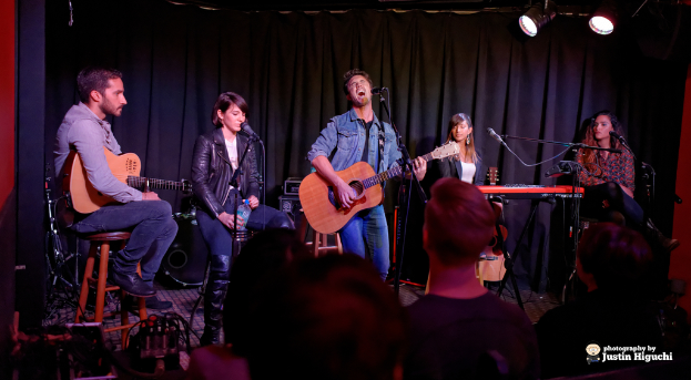 Fünf Personen auf der Bühne, zwei spielen Gitarren, alle haben Mikrofone auf Ständern, mit einem Vorhang und Scheinwerfern im Hintergrund.
