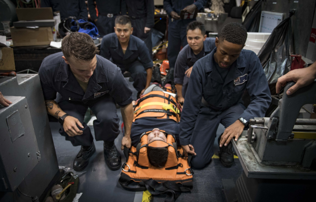 Eine Gruppe von Männern in blauen Uniformen betreut eine Person auf einer Trage in einem chaotischen Raum mit medizinischer Ausrüstung und Kartons, während andere im Hintergrund beobachten.