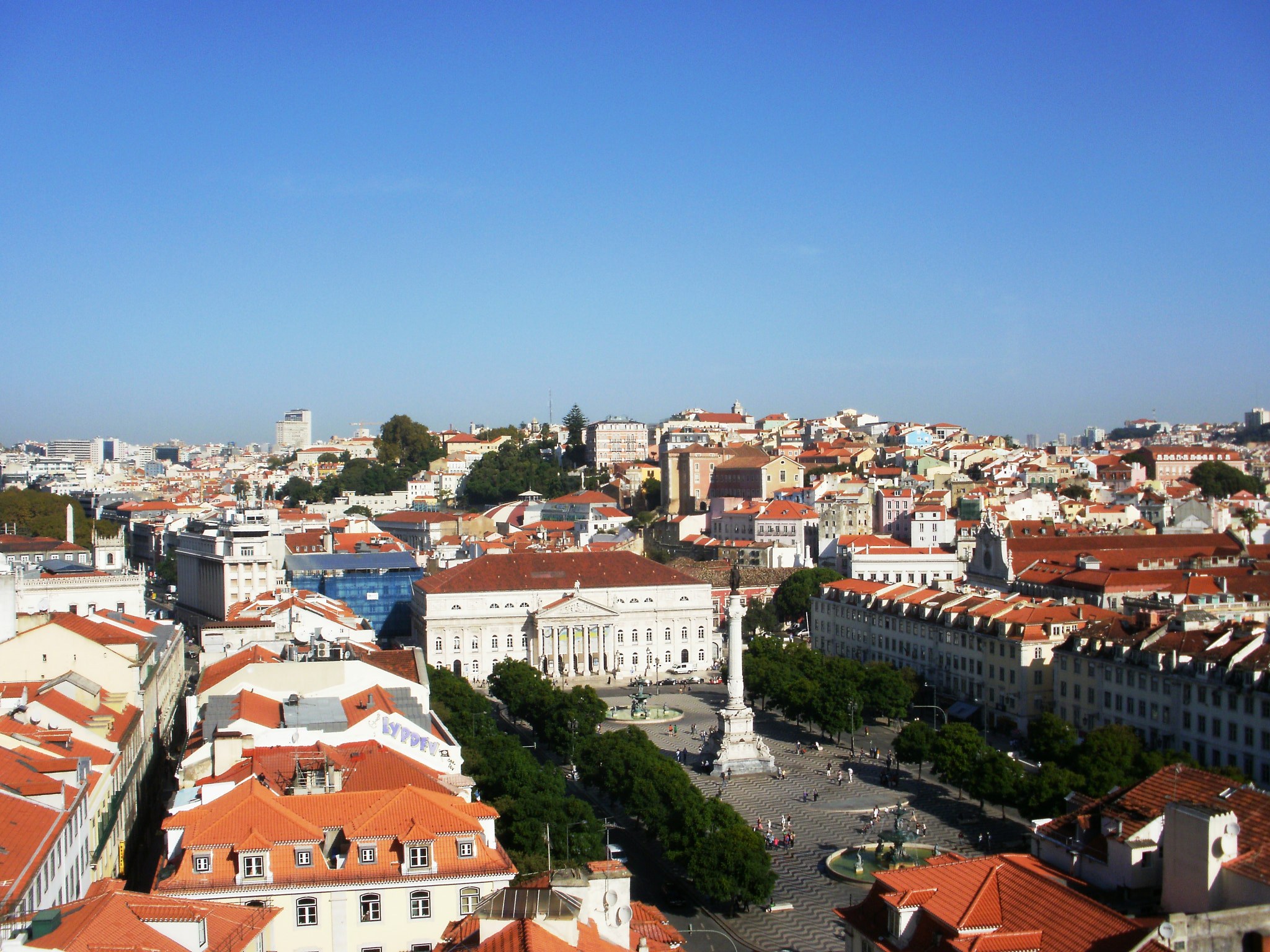 Blick auf Lissabon von einem Hügel mit Gebäuden, Bäumen, einer Statue auf einem Sockel, Menschen auf der Straße und dem Himmel im Hintergrund.