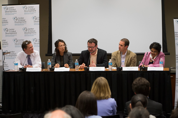Ein Konferenzraum mit einem Tisch, der Namensschilder, Flaschen und ein Mikrofon enthält, fünf Personen, die darum sitzen, ein Podium und ein Banner auf der linken Seite und ein Publikum davor.