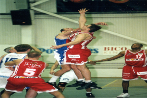 Eine Gruppe von Männern, die Basketball auf einem Court mit einer Wand im Hintergrund spielen, auf der Text und Lautsprecher zu sehen sind, und ein Basketball in der Luft an der Spitze des Bildes.