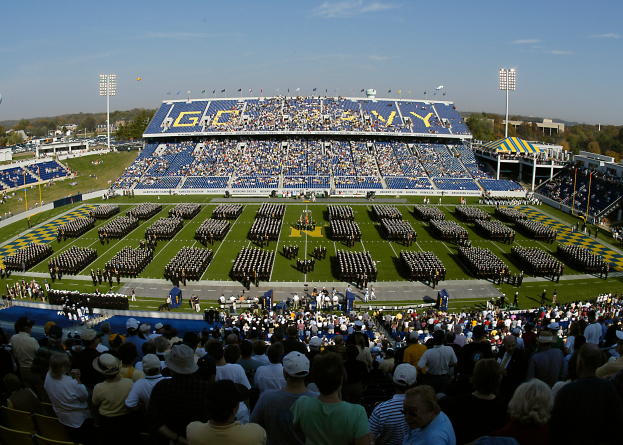 Eine große Menschenmenge in einem Stadion, die ein Fußballspiel sieht, mit einigen Stehenden und anderen Sitzenden auf einem Rasenfeld unter einem klaren blauen Himmel, mit Laternen, Bäumen und Gebäuden im Hintergrund.