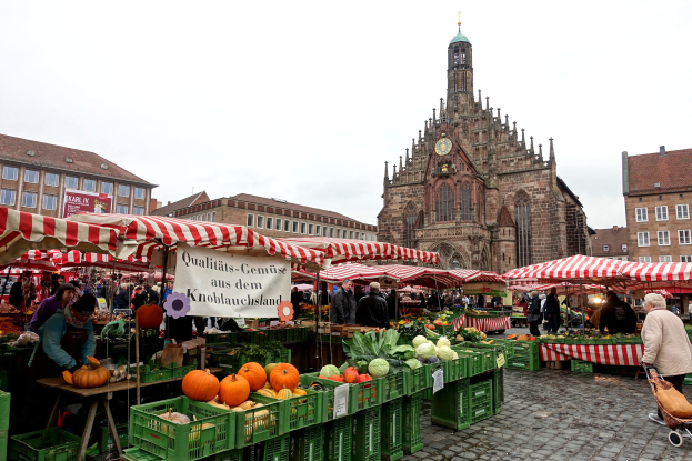 Ein belebter Markt in Nürnberg, Deutschland, mit verschiedenen Früchten und Gemüsen auf dem Markt, Einkaufenden, Zelten und Gebäuden mit einem Uhrenturm im Hintergrund bei klarem Himmel.