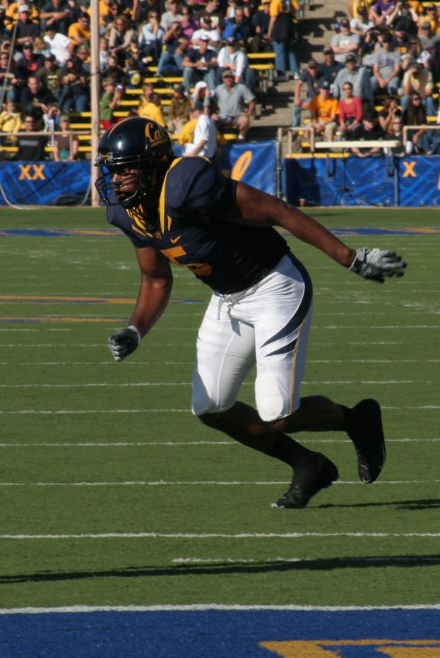 Ein Fußballspieler in einem Sportoutfit, Helm, Handschuhen und Schuhen rennt mit dem Ball auf einem Feld, mit Zuschauern im Hintergrund und Bannern mit Text.