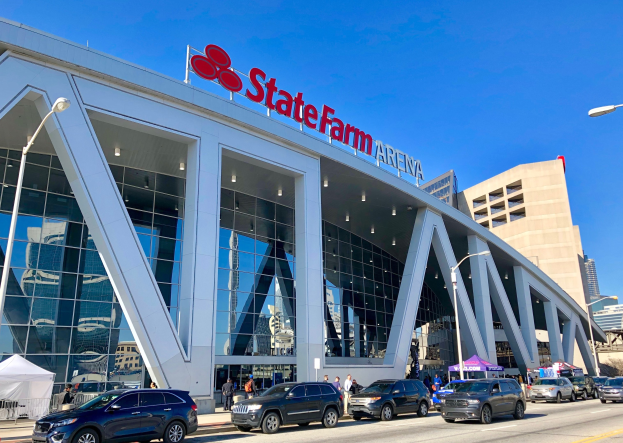 Außenansicht der State Farm Arena in Minneapolis, Minnesota, mit Glaswänden, Säulen, einem Namensschild, Laternen, Fahrzeugen, Fußgängern, Bäumen und einem klaren blauen Himmel.