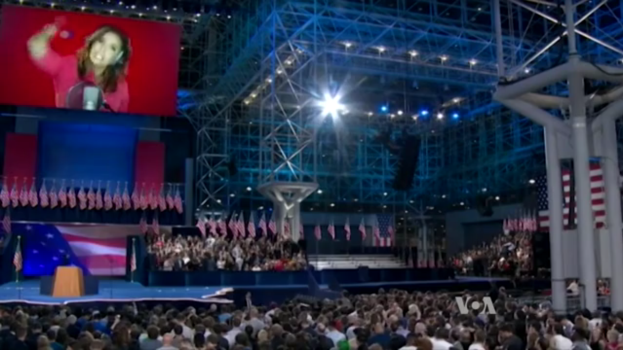 Eine große Menschenmenge sitzt vor einer Bühne mit einem Podium, einem Mikrofon, Flaggen, einem Bildschirm mit einer Frau und Lichtausrüstung, wahrscheinlich bei einer Präsidentenrede.