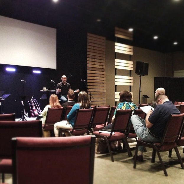 Ein Innenraum mit Frauen auf roten Ledersesseln, ein Mann in einem schwarzen Hemd, der vor ihnen spricht, ein weißer Projektionsschirm, hölzerne Wanddekorationen, ein Lautsprecher auf einem Ständer und eine schwarze Decke mit Scheinwerfern.