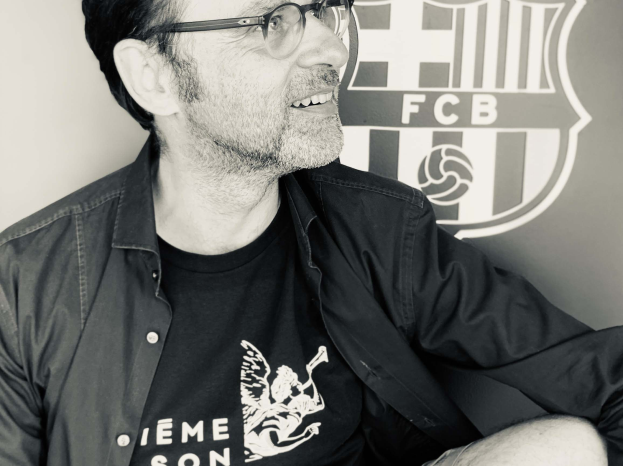 Ein Mann mit Brille und schwarzem Hemd sitzt lächelnd vor einem schwarz-weißen FC Barcelona-Logo an einer Wand.