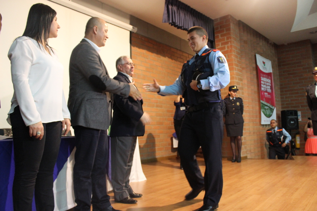 Eine Gruppe von Menschen auf einer Bühne, einer in Uniform mit einer Mütze, eine Frau in einem weißen Hemd, mit einem Banner, Lautsprecher, Vorhängen, Tisch und Wandmonitor im Hintergrund.