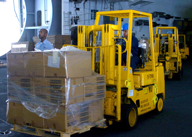 Zwei Männer in blauen Helmen bedienen gelbe Gabelstapler in einem Lagerhaus, das mit Kartons und anderen Gegenständen auf einem Hintergrund mit Rohren und anderen Objekten gefüllt ist.