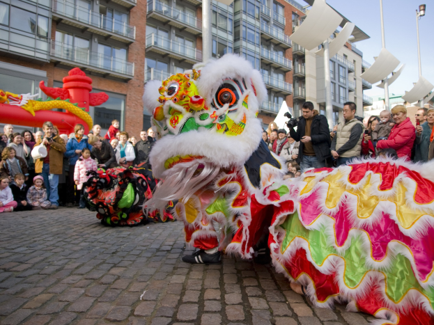 Ein farbenfrohes chinesisches Neujahrsfest in Amsterdam mit einem Löwen tanzen im Vordergrund und einer Menge Menschen drumherum, einige halten Kameras, vor Häusern, Laternenmasten und einem klaren blauen Himmel.